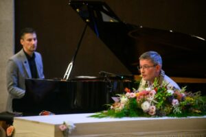 Man spreekt op podium met bloemen, pianist op de achtergrond, vleugelpiano zichtbaar.